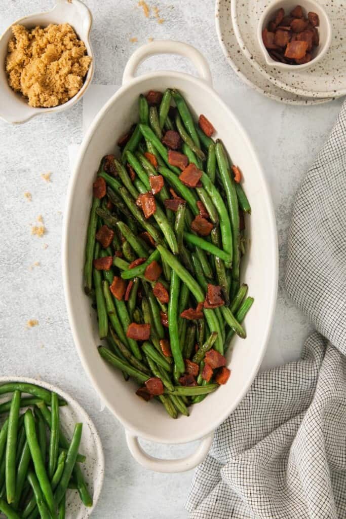 Green beans with bacon in an oblong serving dish surrounded by extra green beans, bacon and brown sugar.