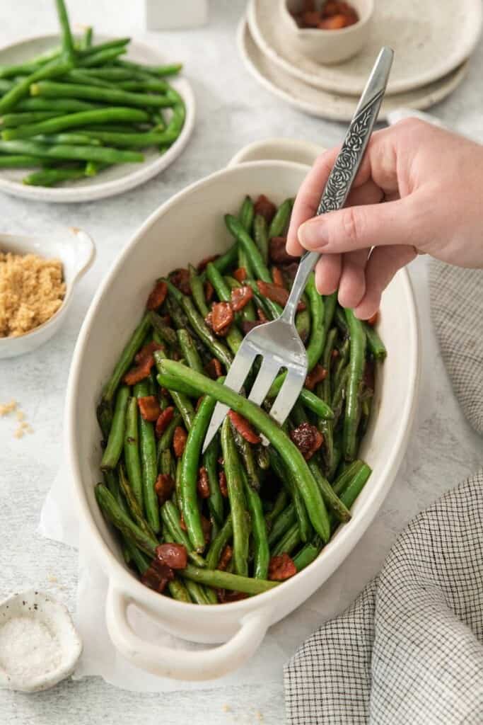 A fork lifting a bite of green beans with bacon from an oblong baking dish.