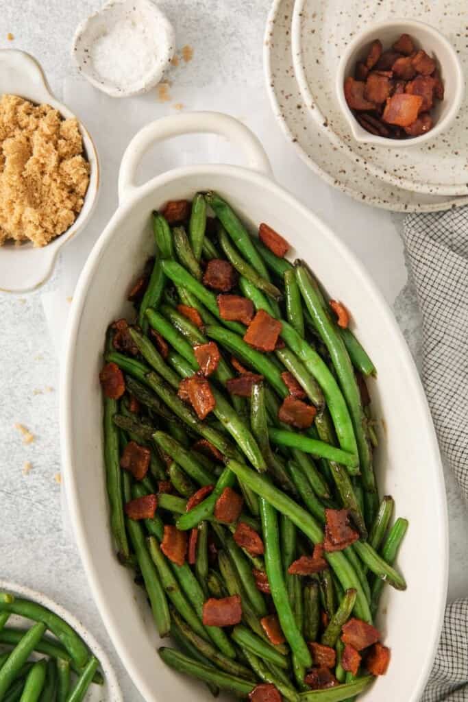 Vertical view of green beans with bacon in an oblong serving dish on a white tablecloth.