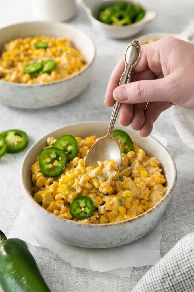 A spoon lifting a bite of jalapeno cream corn from a white bowl.