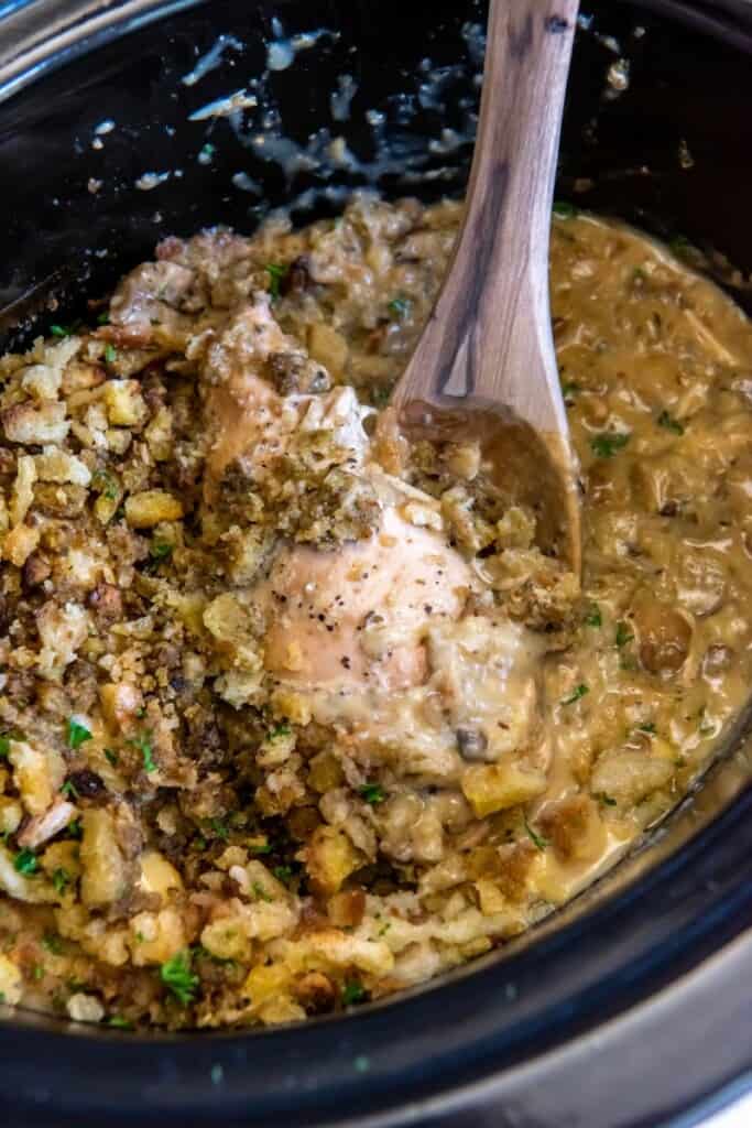 A wooden spoon lifting chicken and stuffing in a crock pot.