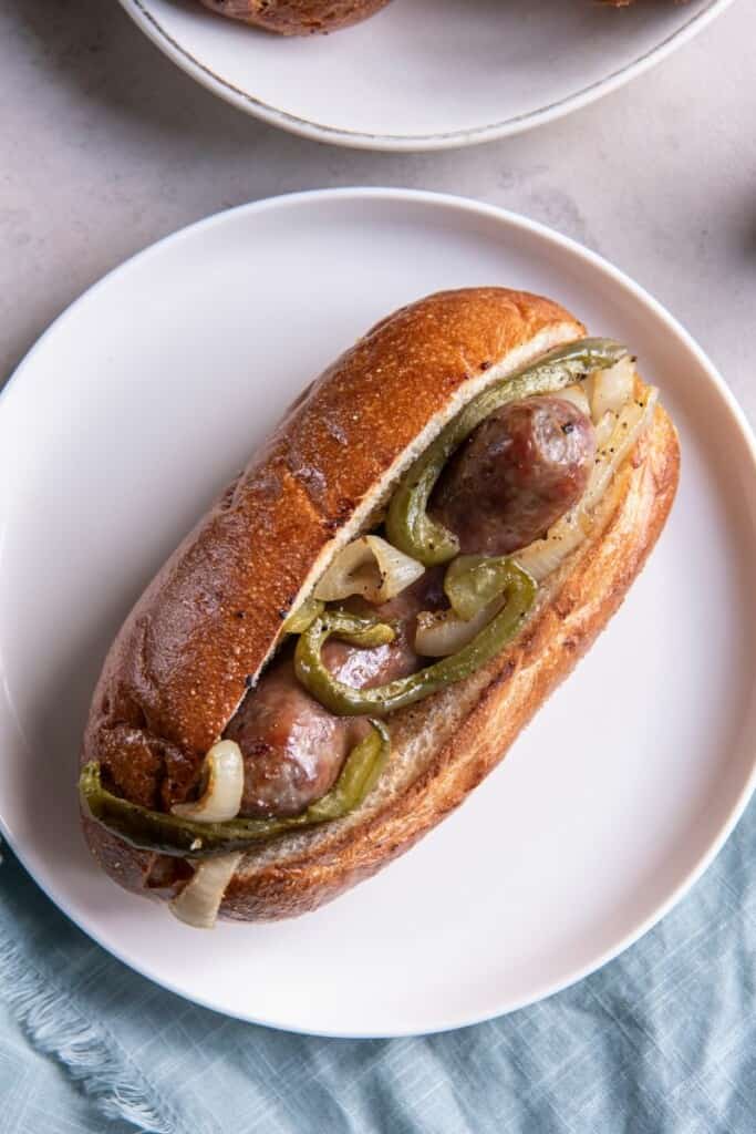 A baked brat in a bun with onions and peppers on a white plate.