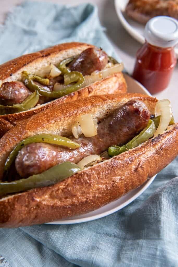 Two brats in buns with onions and peppers on a white plate with a blue background.