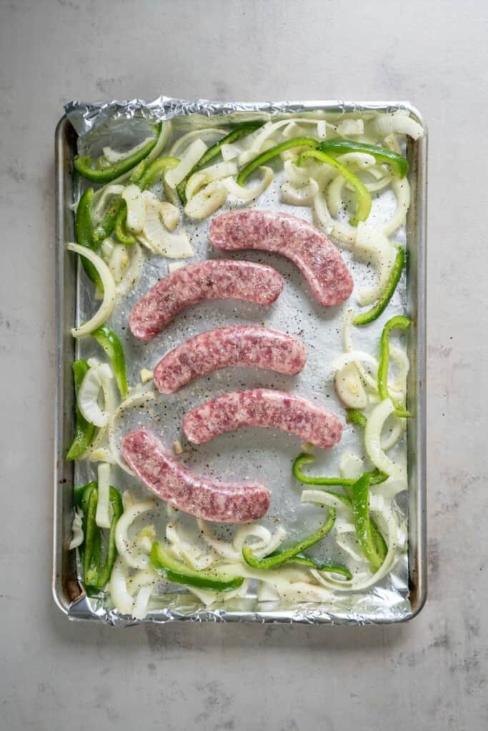 Brats with seasoned onions and peppers on a foil lined baking sheet.