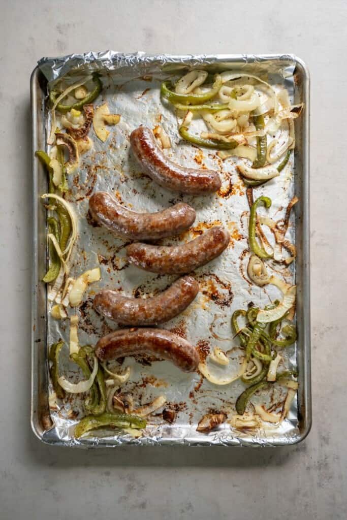 Baked brats, onions and peppers on a foil lined baking sheet.