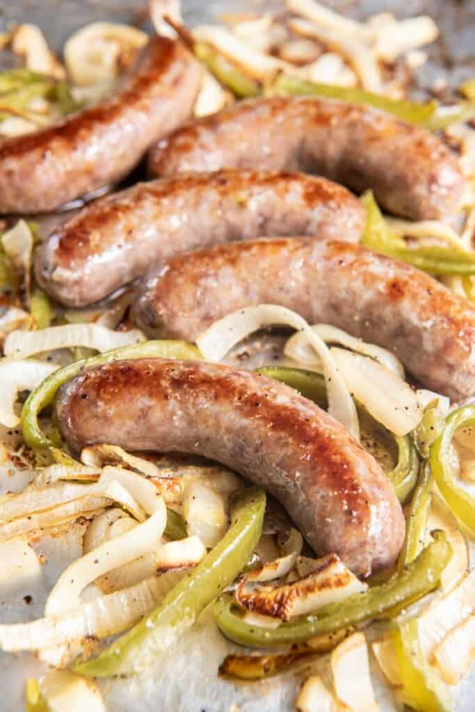 Closeup view of baked brats, onions and peppers on a foil lined baking sheet.