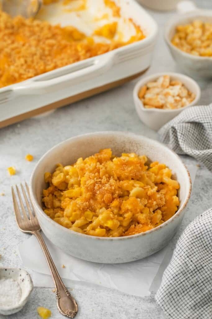 A bowl of corn macaroni casserole next to a fork with the casserole dish in the background.