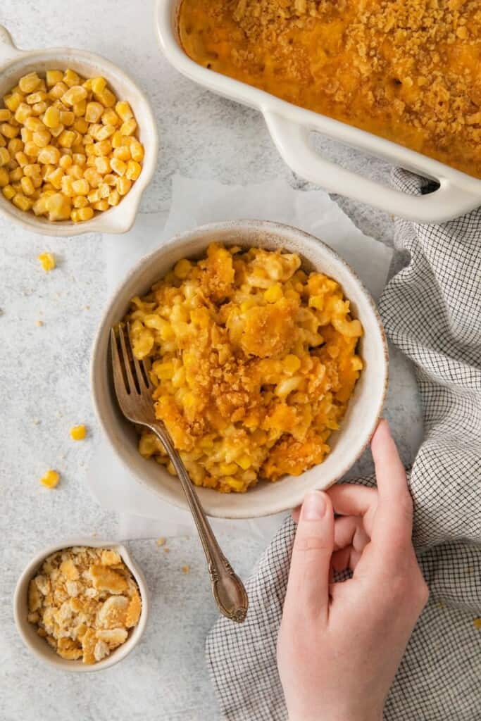 A bowl of macaroni corn casserole surrounded by cheese and the casserole dish.