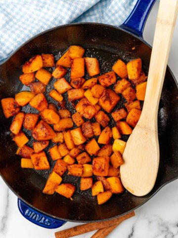 Diced butternut squash that has been sauteed in a skillet with a wooden spoon.