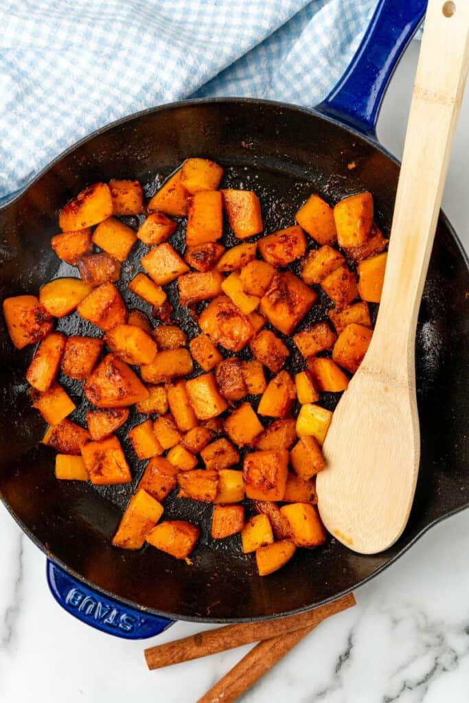 Diced butternut squash that has been sauteed in a skillet with a wooden spoon.
