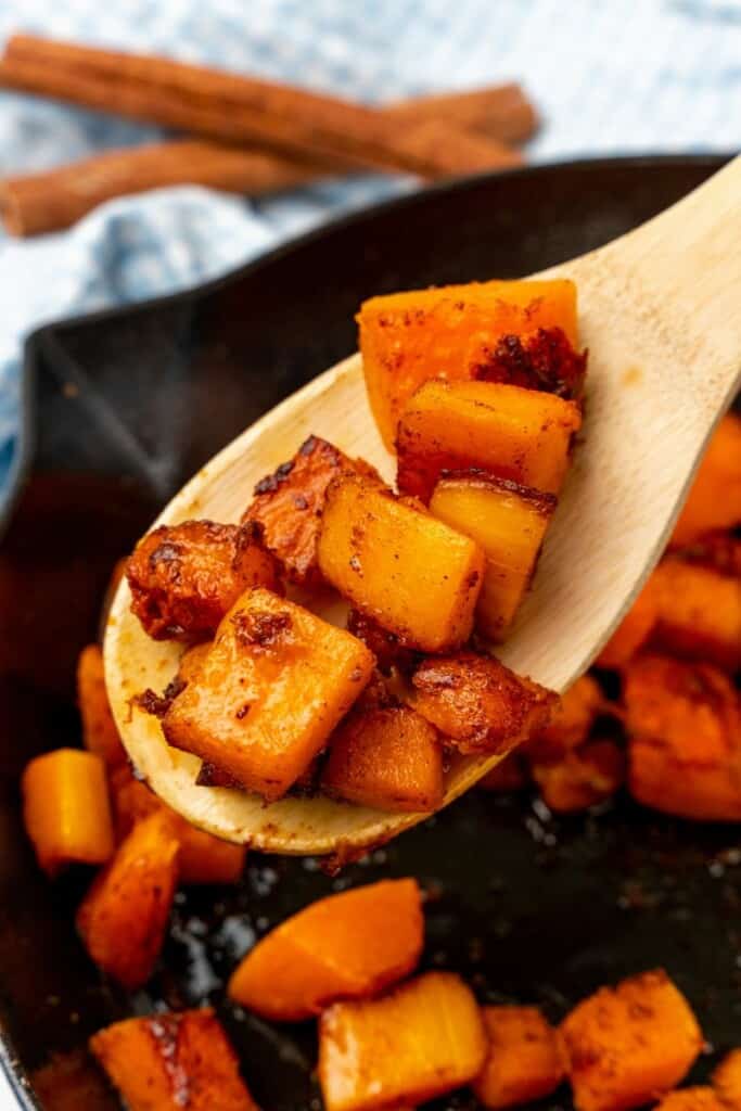 Wooden spoon holding a scoop of pan fried butternut squash over a skillet.