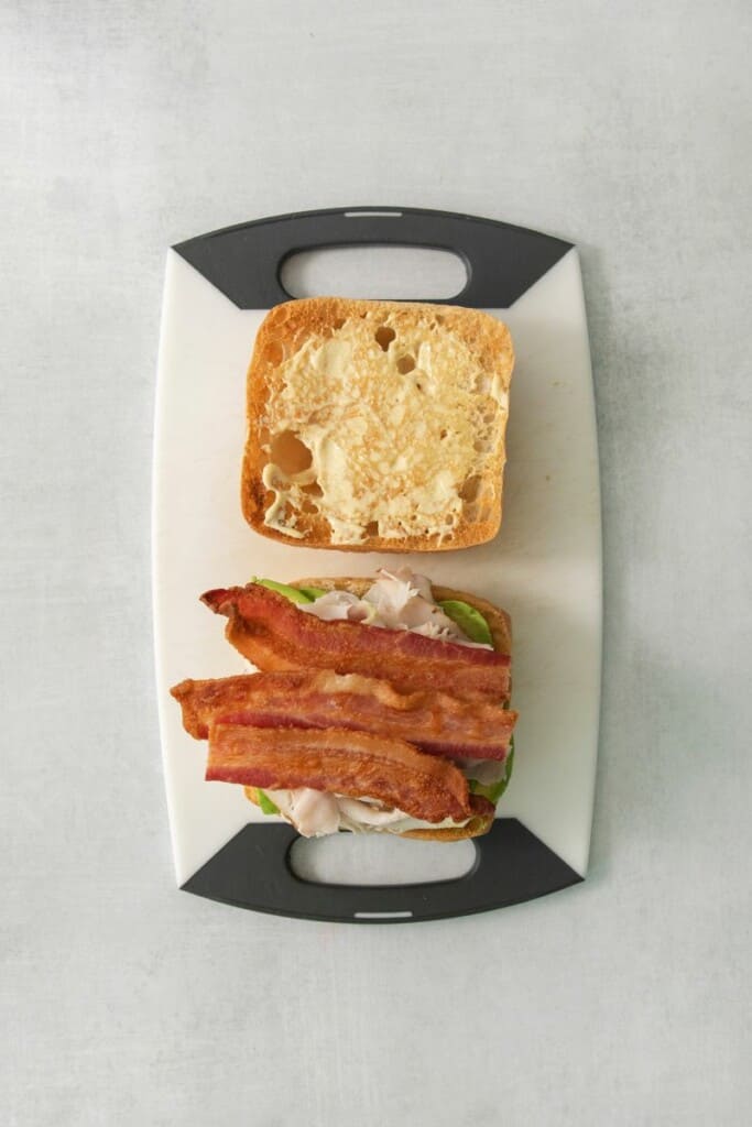Toasted sourdough, avocado, turkey, and bacon on a cutting board.