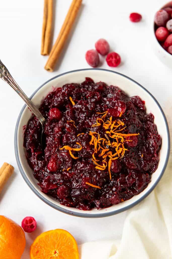 Cranberry orange sauce in a bowl with orange zest on top, with a spoon inside of it, and cinnamon sticks and oranges around it.