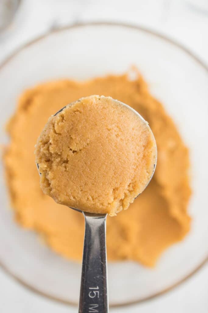 A measuring spoon holding a ball of peanut butter cookie dough over the mixing bowl of additional dough.