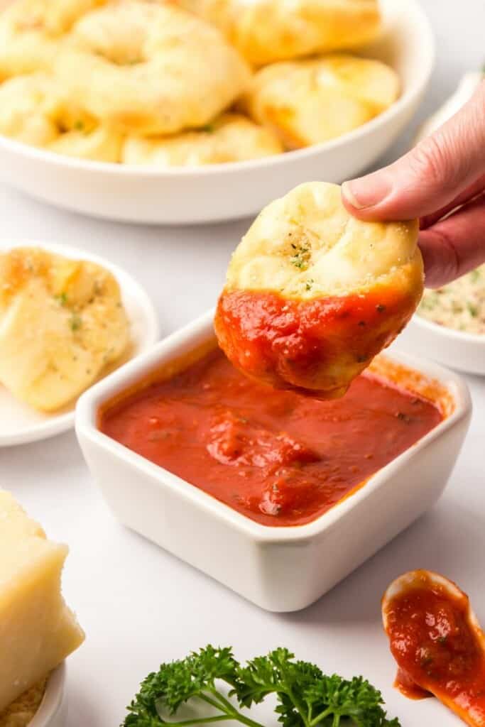 A garlic knot dipped into marinara sauce with a plate of additional garlic knots in the background.