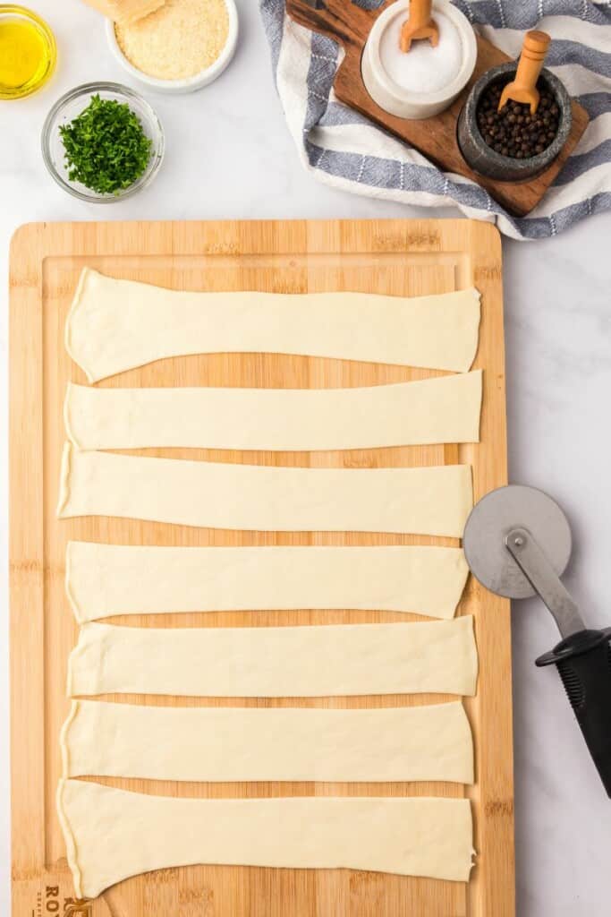 Rolling out dough on a wooden cutting board to be cut into strips with a pizza cutter.