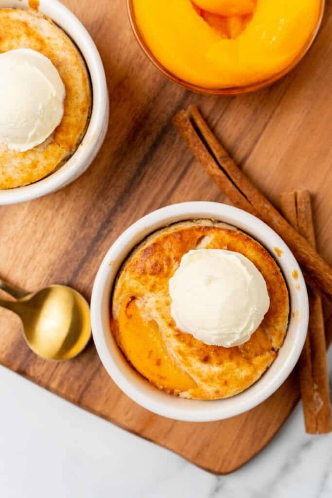 Two individual peach cobblers with whipped cream on top resting on a wooden cutting board.