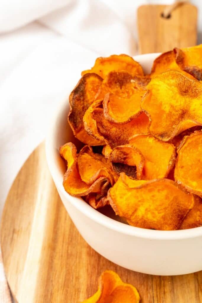 A white bowl of sweet potato chips on a wooden cutting board.