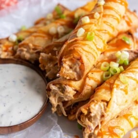 A stack of prepared buffalo chicken taquitos with green onions on top and dipping sauce next to the stack,