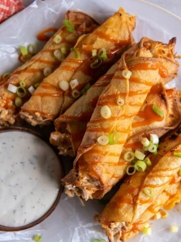 A stack of prepared taquitos topped with green onions and drizzled with buffalo sauce.