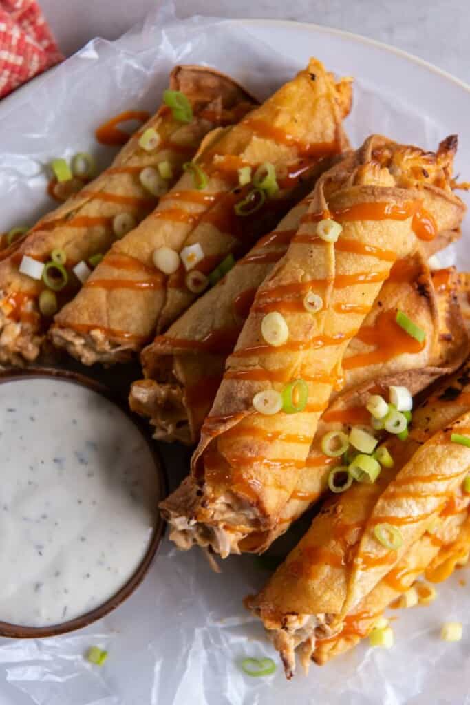 A stack of prepared taquitos topped with green onions and drizzled with buffalo sauce.