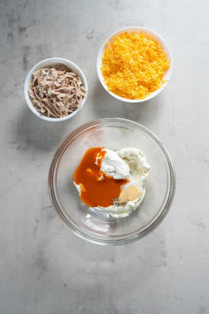 A bowl of ingredients to combine for buffalo sauce, a bowl of shredded chicken and a bowl of cheese.