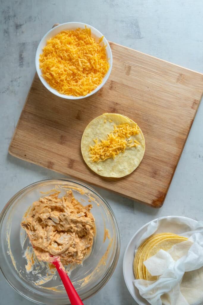 A bowl of shredded chicken coated in buffalo sauce, a bowl of cheese, shredded cheese on a tortilla resting on a wooden cutting board.