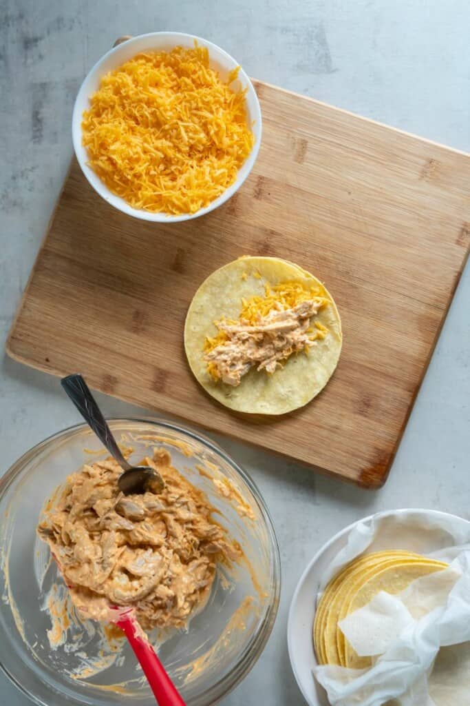Adding buffalo chicken to cheese on a tortilla resting on a wooden cutting board. A small bowl of cheese. A small bowl of buffalo chicken.