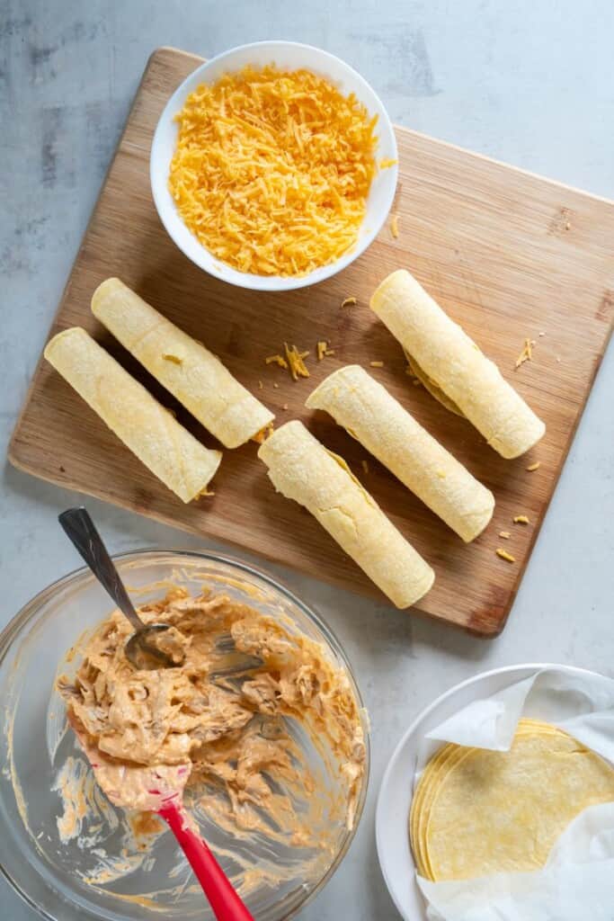 Five rolled uncooked taquitos on a wooden cutting board next to a bowl of cheese and a bowl of buffalo chicken.