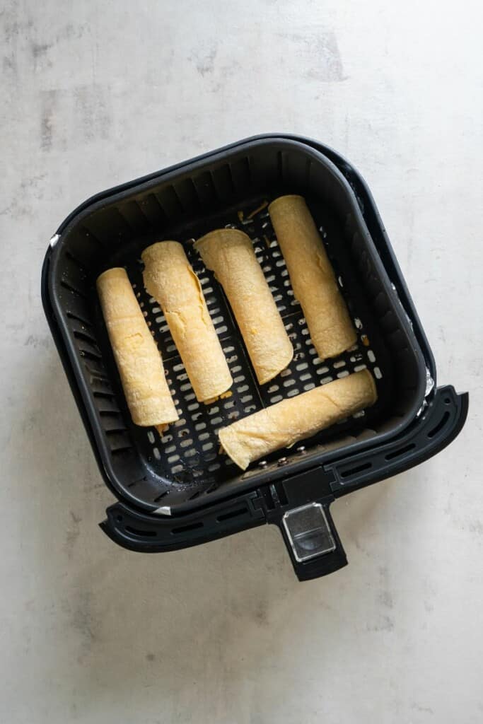Five uncooked taquitos resting in a black air fryer basket.