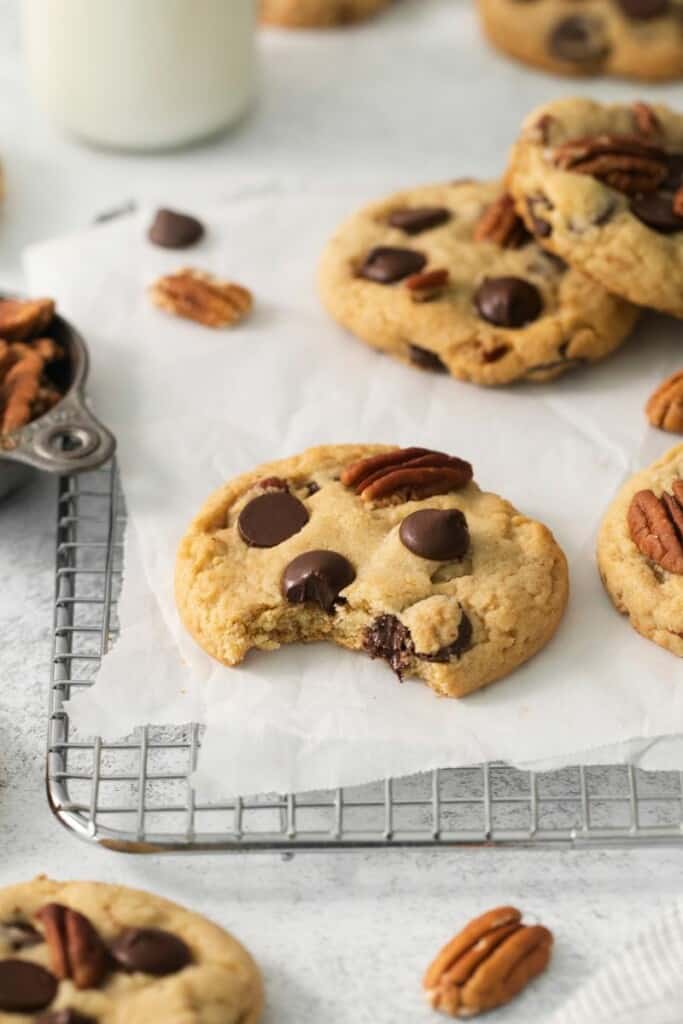 Baked pecan chocolate chip cookies on a parchment lined cooling rack, one cookie has a bite removed.