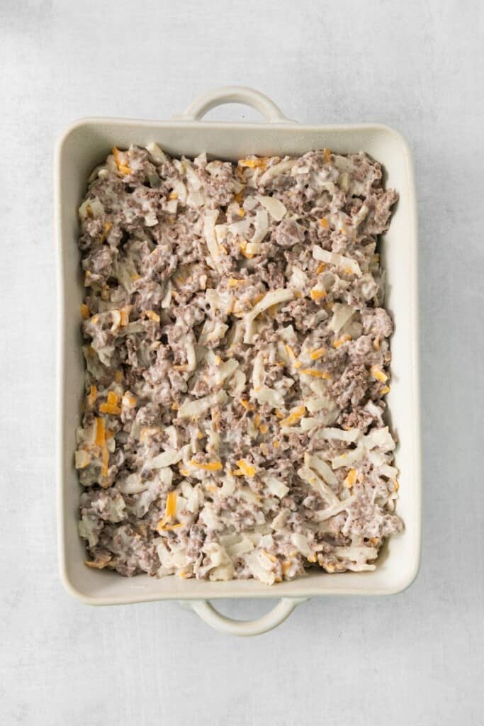 Spreading ingredients for hamburger hash brown casserole in a baking dish.