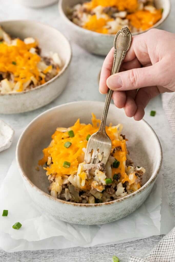 A fork holding a bite of hamburger hash brown casserole over a bowl containing one serving of this tasty dish.