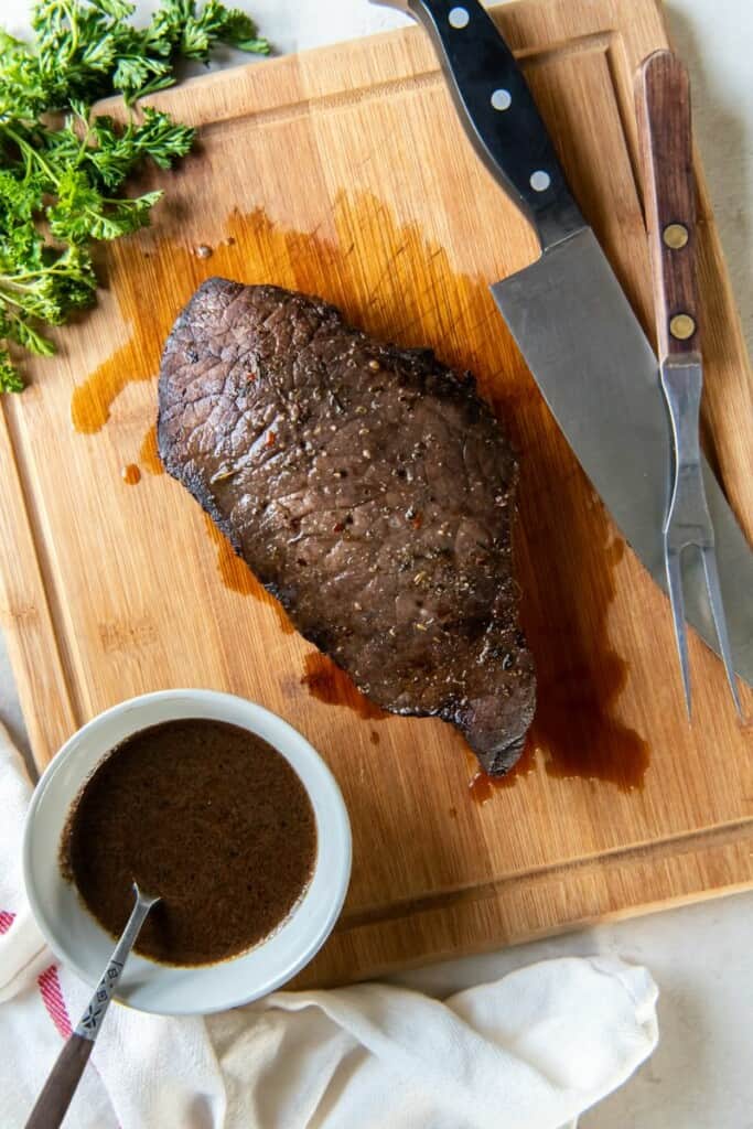Prepared london broil resting on a wooden cutting board next to a chef's knife and saucepan of marinade.