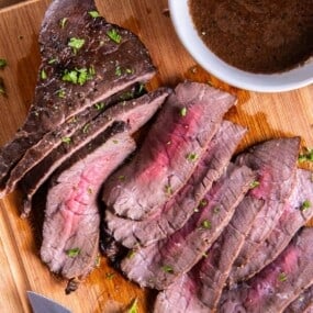 Slices of london broil on a wooden cutting board with a small bowl of marinade.