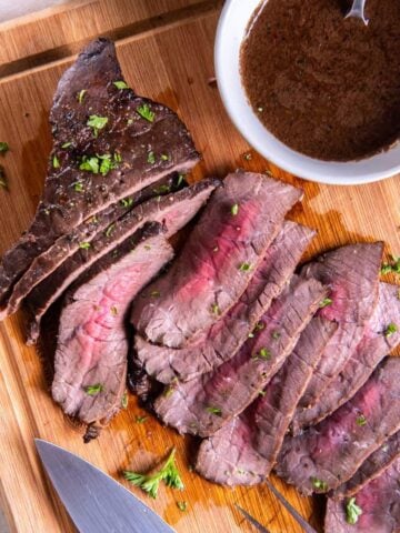 Slices of london broil on a wooden cutting board with a small bowl of marinade.