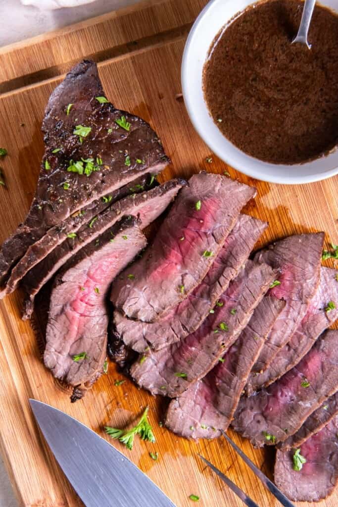 Slices of london broil on a wooden cutting board with a small bowl of marinade.