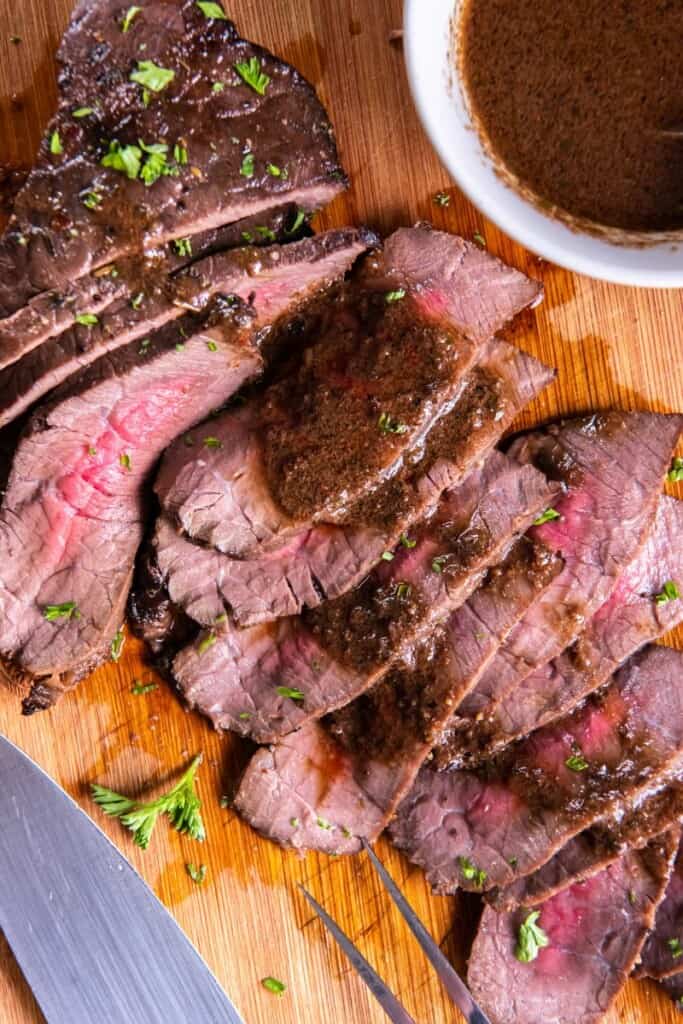 Closeup view of slices of london broil on a wooden cutting board next to a small saucepan of marinade.