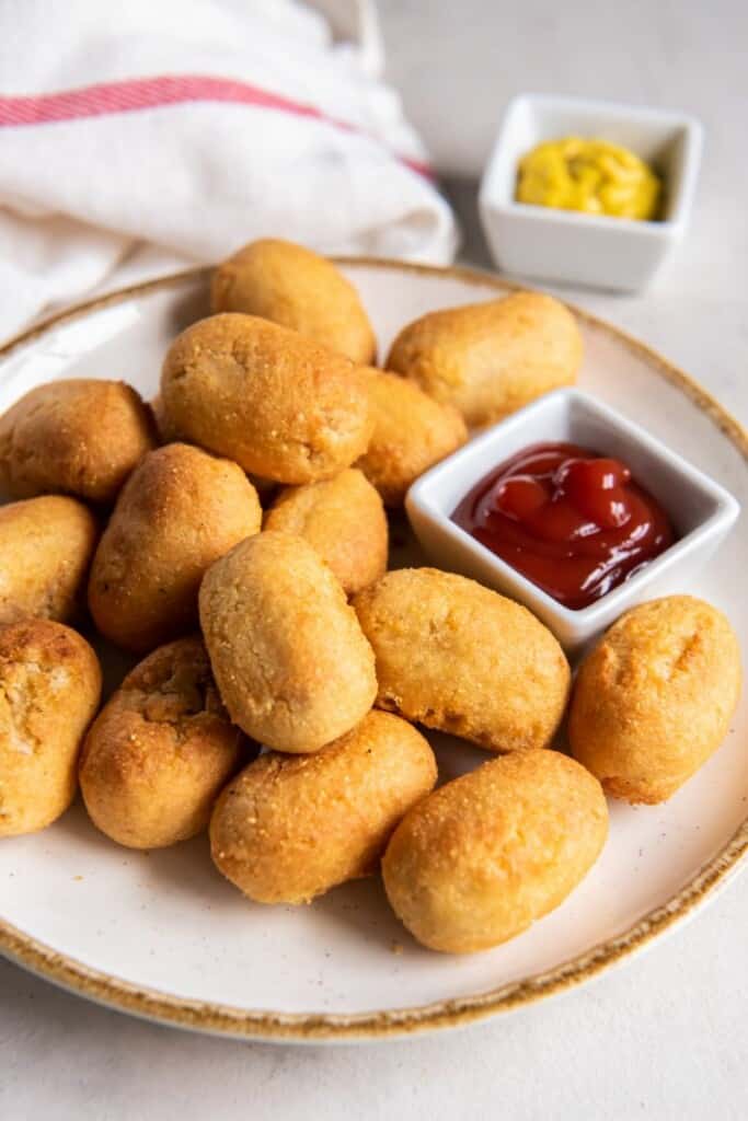 Miniature corn dogs on a white plate with ketchup and mustard for dipping.