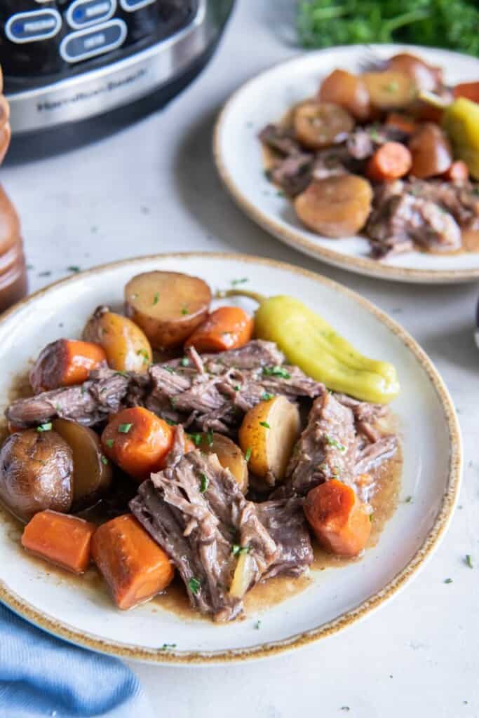 A serving of Mississippi Pot Roast and vegetables plated in front of slow cooker.