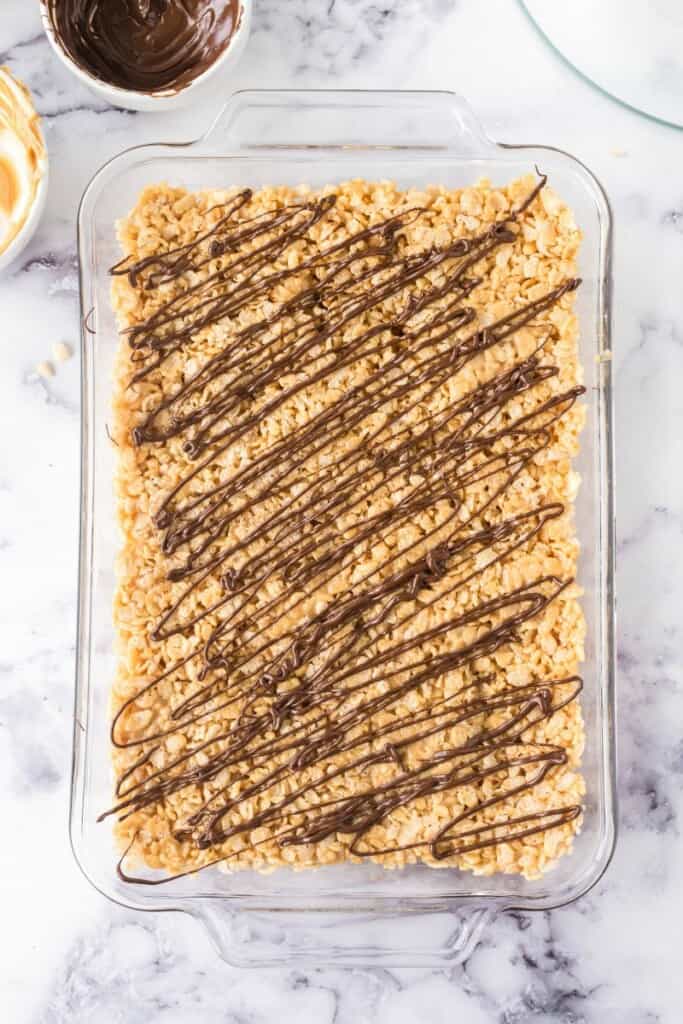 Drizzling melted chocolate over rice krispie mixture in a clear baking dish.