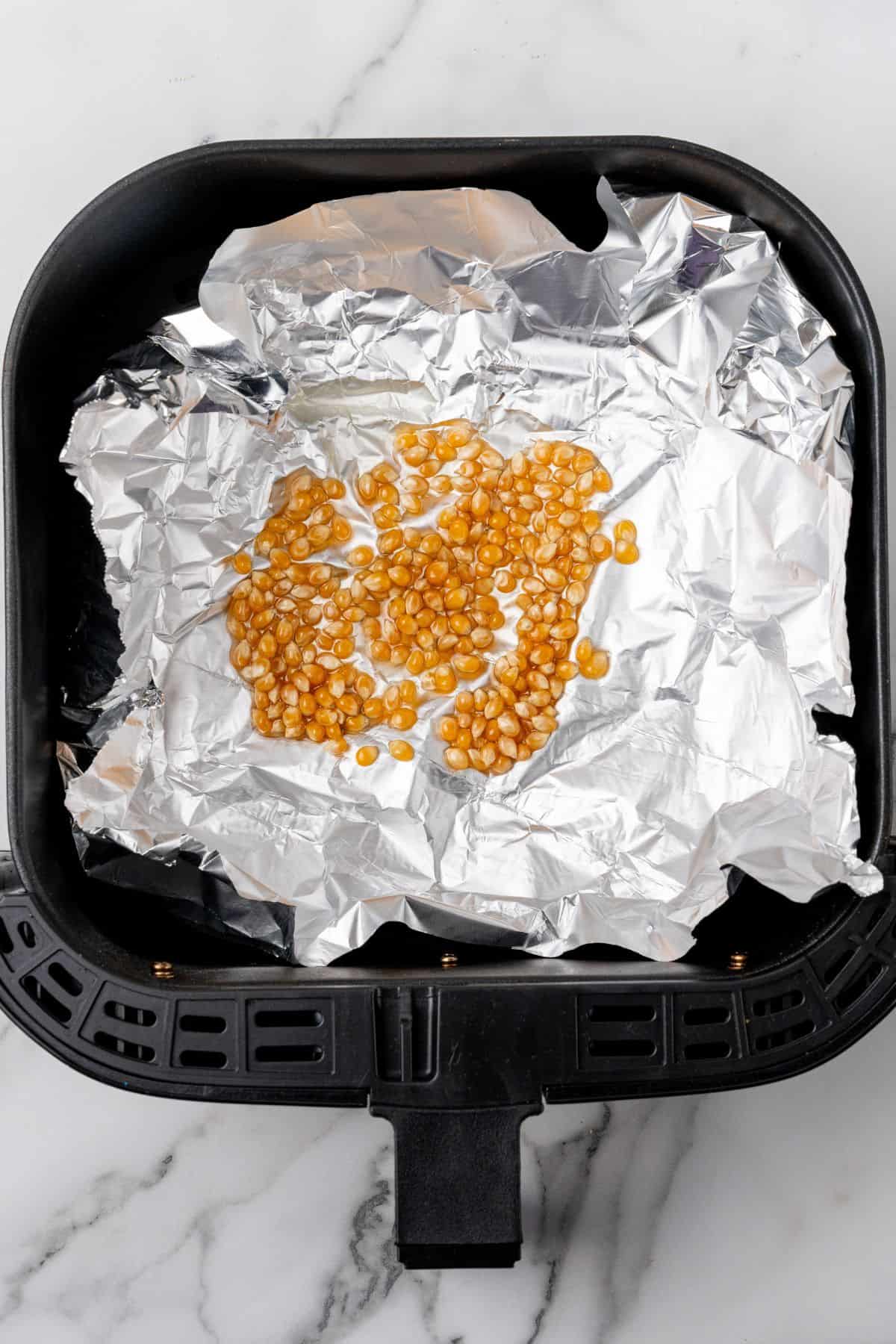 Popcorn kernels in an air fryer basket with foil underneath.
