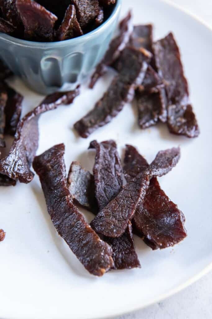 Zoomed in view of beef jerky slices on a white plate with a small blue bowl of additional pieces in the background.