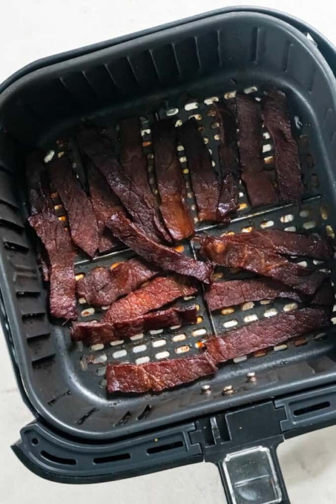 A closeup view of beef jerky slices in a black air fryer basket.