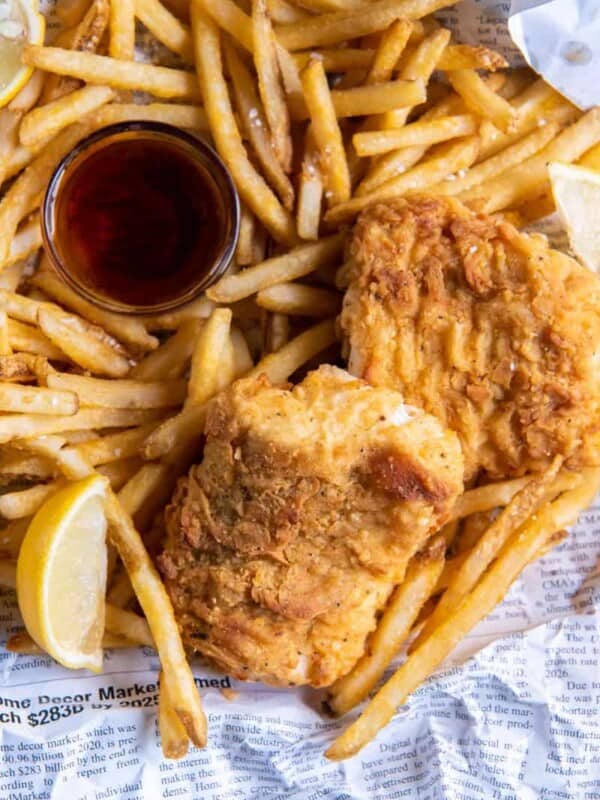 A basket of prepared fish and chips with lemon wedges and dipping sauce.