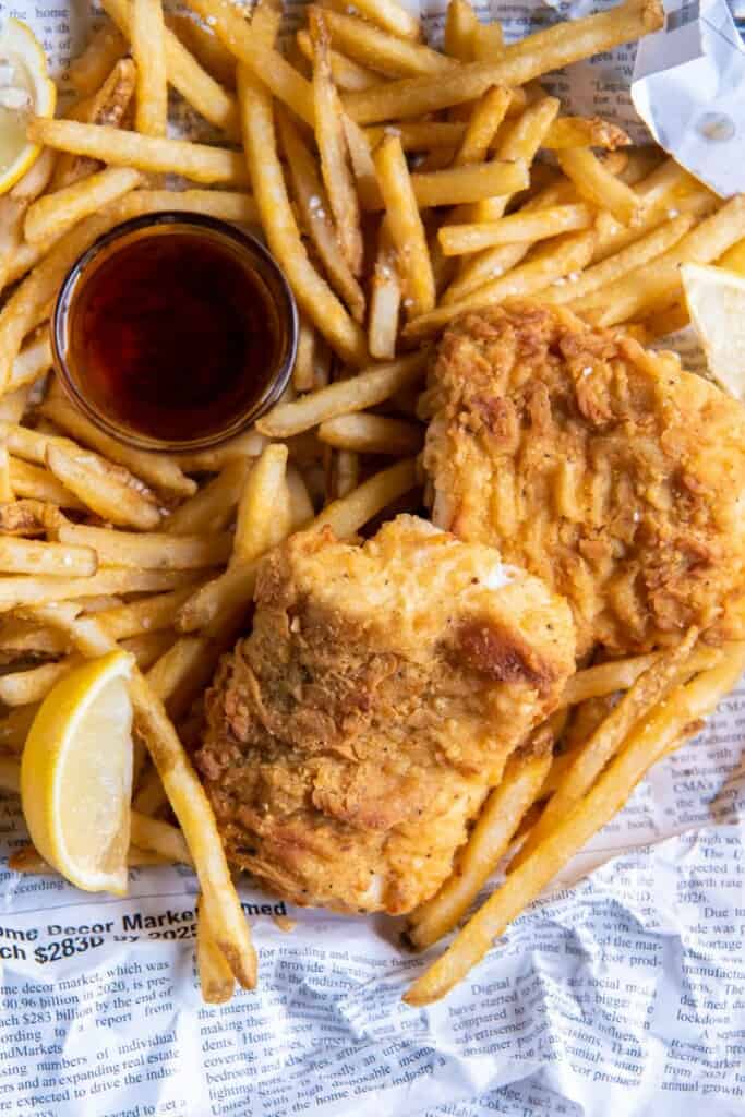 A basket of prepared fish and chips with lemon wedges and dipping sauce.