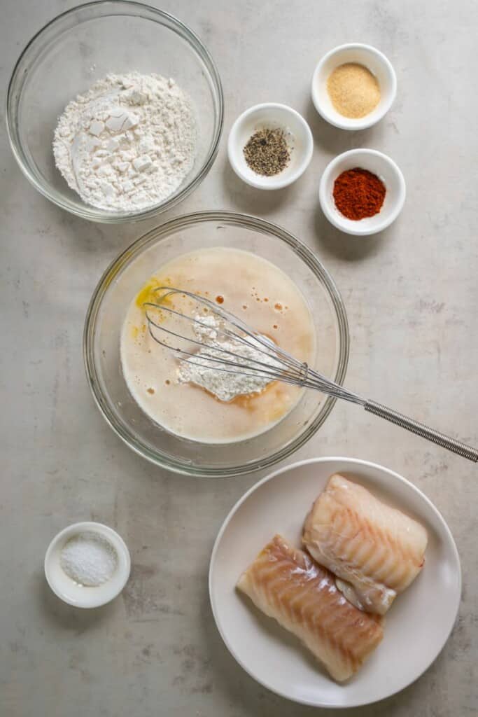A plate of cod, several small bowls with individual ingredients to prepare fish batter, one larger bowl with the fish batter ingredients.