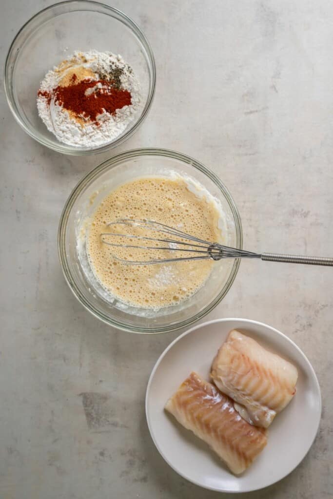 A bowl of seasoned flour, a bowl of fish batter, a plate with two cod filets.