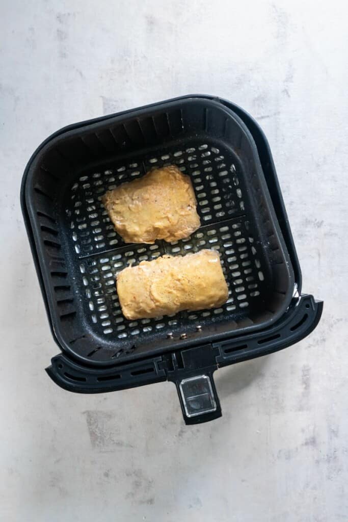 Two cod filets resting in a black air fryer basket after cooking for the first 12 minutes.