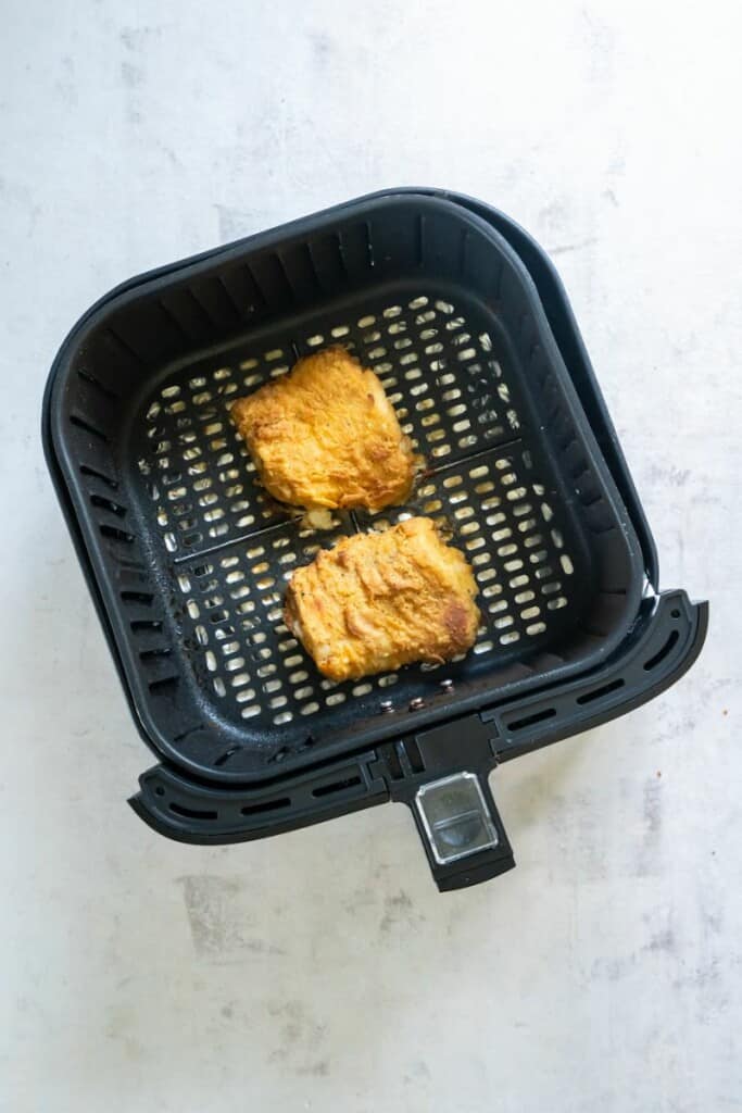 Two cod fish filets resting in a black air fryer basket after cooking the full amount of required time.